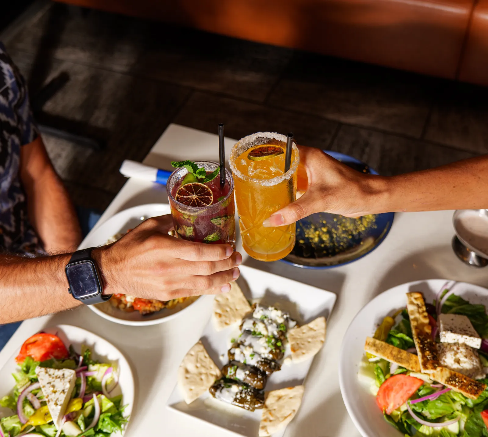 Two people toasting cocktails over a Greek dinner spread