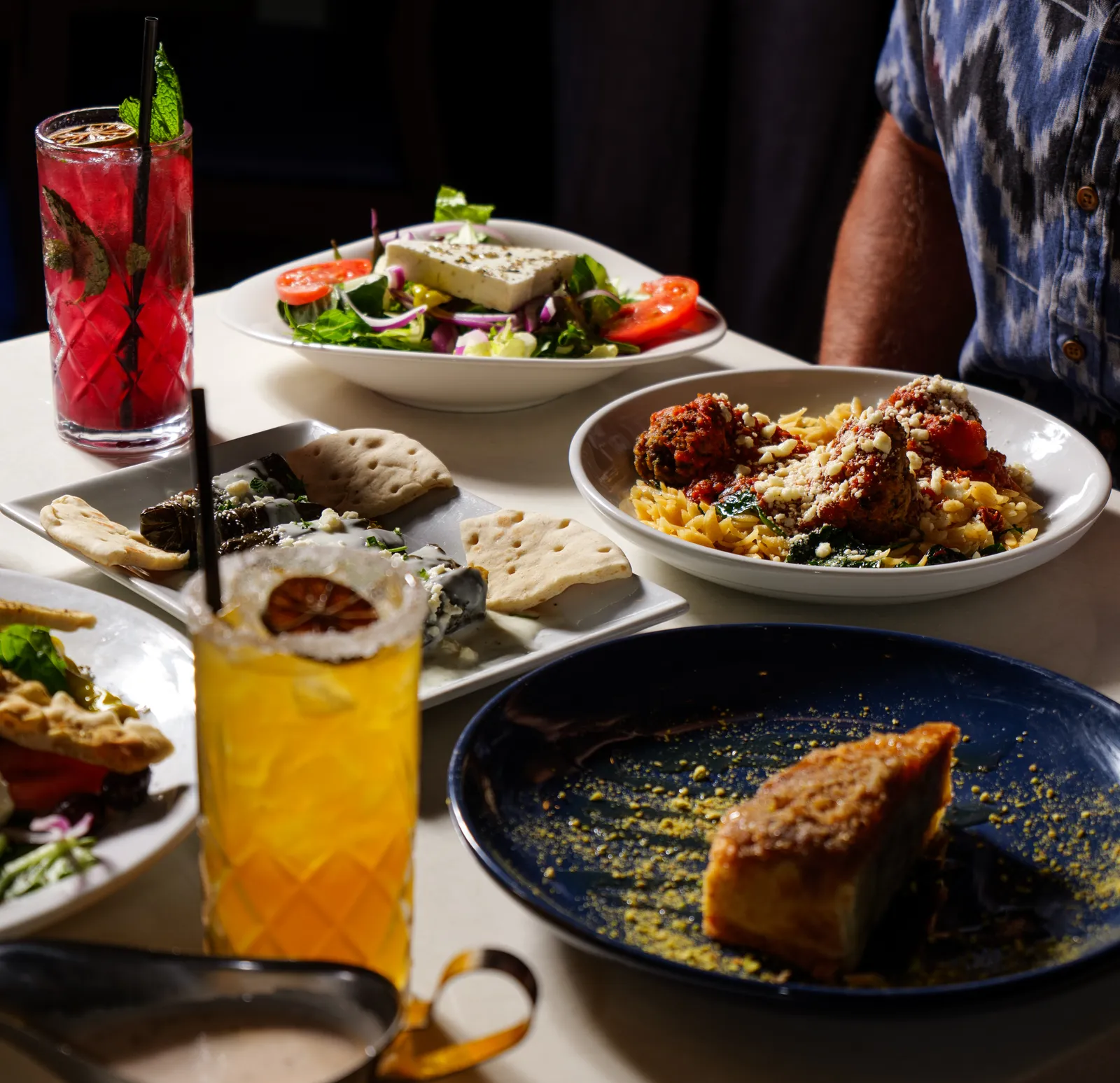 Greek dinner spread with salad, pasta, flatbread, dessert and an orange cocktail